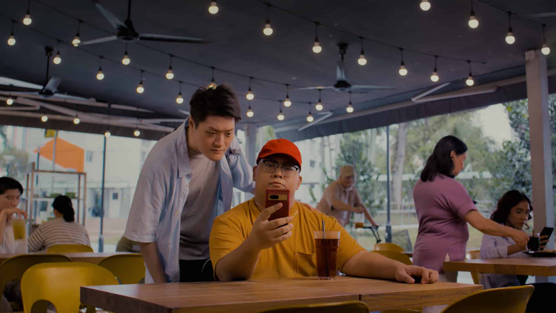 “Two young men at an outdoor café, one seated in a red cap looking at his phone while the other leans over to view the screen, with string lights and other patrons in the background.